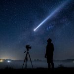 Nuit d’août : un photographe en silhouette sur un trépied, avec une traînée lumineuse d’étoile filante au-dessus d’un paysage sombre.