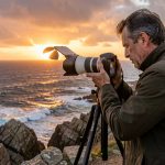 Un photographe observe une éclipse solaire partielle au-dessus d’une côte française au coucher du soleil, avec un appareil hybride monté sur trépied et équipé d’un filtre solaire.