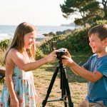 Une jeune fille et un jeune garçon réalisent un petit film de vacances avec une caméra posée sur un trépied, dans un camping ensoleillé au bord de la mer.
