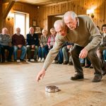 Un homme âgé lance un palet dans une salle associative chaleureuse à Availles-Limouzine, tandis qu’un public composé de générations différentes observe la partie.